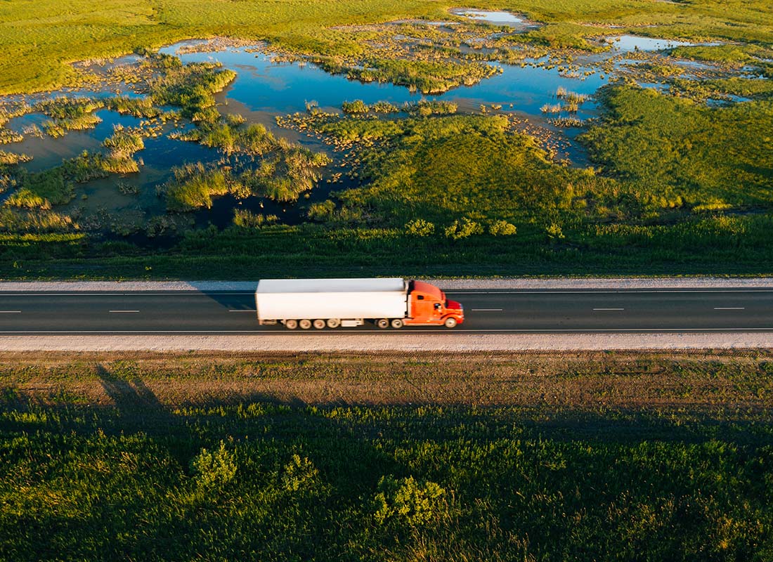We Are Independent - Aerial View of a Truck with a Trailer Driving Down a Road at Sunset Next to a Lake and Fields with Green Grass